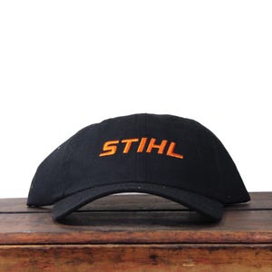 Pode incluir: Boné de beisebol preto com a palavra "STIHL" bordada em laranja na frente. O boné está sobre uma superfície de madeira. O fundo é branco.