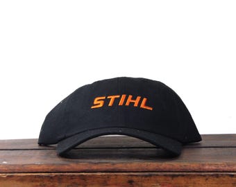 Op de afbeelding: Zwarte baseball cap met het woord "STIHL" in oranje geborduurd op de voorkant. De cap ligt op een houten oppervlak. De achtergrond is wit.