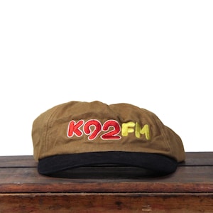 Peut inclure: Casquette plate marron avec une visière noire. Le chapeau porte le texte brodé "KQ2FM" en rouge et jaune. Le chapeau est posé sur une surface en bois.