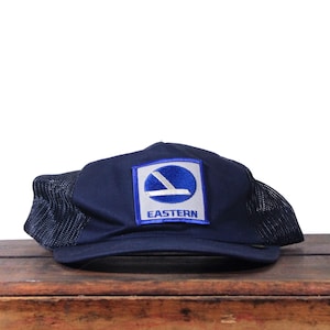 May include: A navy blue trucker hat with a mesh back and a curved brim. The front features a square patch with the word "EASTERN" and a white and blue logo. The hat is sitting on a wooden surface.