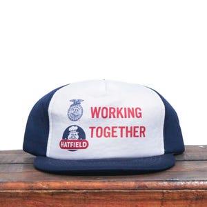 K&ouml;nnte beinhalten: Eine wei&szlig;e und blaue Baseballkappe mit einem dunkelblauen Schirm. Die Vorderseite der Kappe hat einen wei&szlig;en Hintergrund mit dem Text "WORKING TOGETHER" in roten Buchstaben. Unter dem Text befindet sich ein Logo mit einem Schwein und dem Text "HATFIELD".