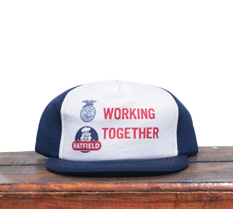 K&ouml;nnte beinhalten: Eine wei&szlig;e und blaue Baseballkappe mit einem dunkelblauen Schirm. Die Vorderseite der Kappe hat einen wei&szlig;en Hintergrund mit dem Text "WORKING TOGETHER" in roten Buchstaben. Unter dem Text befindet sich ein Logo mit einem Schwein und dem Text "HATFIELD".