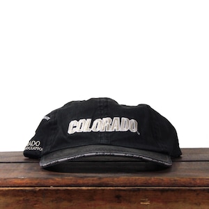 Puede incluir: Gorra de béisbol negra con la palabra "COLORADO" bordada en blanco en la parte delantera. La gorra tiene una visera curva y una correa ajustable. La gorra está sobre una superficie de madera.