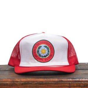 Könnte beinhalten: Eine rot-weiße Trucker-Kappe mit einem weißen Frontpanel, auf dem das Siegel der Cherokee Nation abgebildet ist. Das Siegel ist ein Kreis mit einem grünen Kranz, einem gelben Stern und dem Text "SEAL OF THE CHEROKEE NATION" um den Außenrand. Der Text "JULY 1839" und "SEP. 6, 1839" befindet sich unter dem Kranz.