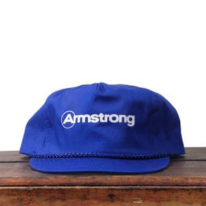Puede incluir: Gorra de béisbol azul real con la palabra "Armstrong" en blanco dentro de un círculo blanco. La gorra tiene una visera curva y un detalle de cuerda a lo largo del borde de la visera.