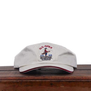 May include: An off-white baseball cap featuring an embroidered U.S. Open 2009 Bethpage Black logo with a graphic of a golfer carrying a bag. The cap has a curved brim with a thin red accent line along the edge and embroidered eyelets on top.