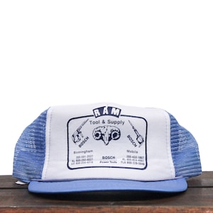 Op de afbeelding: Een blauwe en witte mesh trucker-pet met het logo "RAM Tool & Supply" met een rammenkop en de tekst "Bosch Power Tools" met telefoonnummers voor de locaties Birmingham en Mobile.