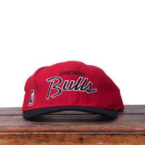 Op de afbeelding: Een rode Chicago Bulls baseball cap met een zwarte klep. De cap heeft de teamnaam in witte letters met een zwarte omlijning en het woord "Chicago" erboven. Het NBA-logo staat op de zijkant.