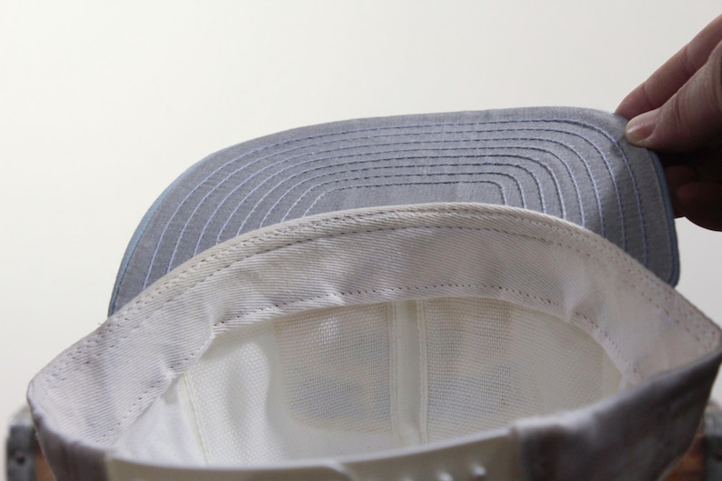 May include: A light gray baseball cap with a white interior and a gray brim. The brim has horizontal stitching. The cap is being held up by a hand. The cap is a classic style.