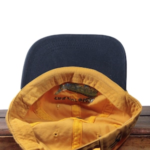 K&ouml;nnte beinhalten: Baseballkappe mit marineblauem Schirm und gelbem Innenraum. Der Innenraum hat eine kleine Grafik und Text. Die Kappe liegt auf einer Holzoberfl&auml;che.