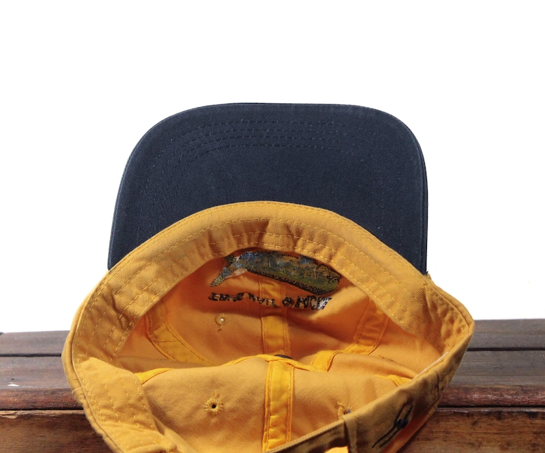 K&ouml;nnte beinhalten: Baseballkappe mit marineblauem Schirm und gelbem Innenraum. Der Innenraum hat eine kleine Grafik und Text. Die Kappe liegt auf einer Holzoberfl&auml;che.