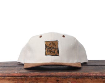 May include: A cream-colored baseball cap with a brown brim and a square patch. The patch features the words "WILL GOLF 4 FOOD" in a brown, textured font. Two small brown dots are on the top of the cap.