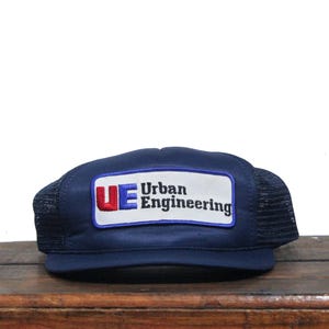 Puede incluir: Una gorra de camionero azul marino con una malla negra en la parte posterior. La gorra tiene un parche blanco con el texto "UE Urban Engineering" en letras azules y rojas.