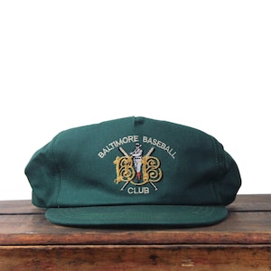 May include: A dark green baseball cap with a flat brim. The cap features an embroidered logo with the words "Baltimore Baseball Club" and a baseball player illustration in gold, white, and red.