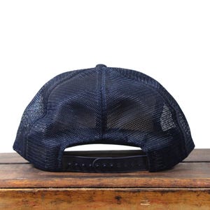 May include: Navy blue trucker hat with a mesh back and adjustable snap closure. The hat sits on a wooden surface. The hat's design includes a curved brim and a structured front panel. This hat is a classic accessory.
