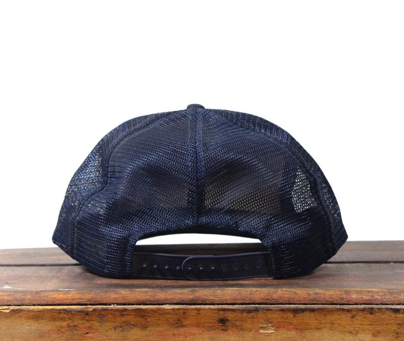 May include: Navy blue trucker hat with a mesh back and adjustable snap closure. The hat sits on a wooden surface. The hat's design includes a curved brim and a structured front panel. This hat is a classic accessory.