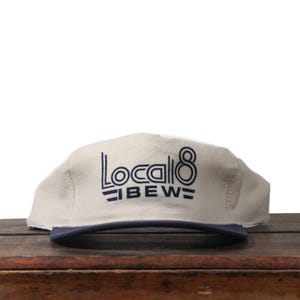 Könnte beinhalten: Beige Kappe mit marineblauem Schirm. Auf der Kappe steht in Marineblau der Text "Local 8 IBEW". Die Mütze liegt auf einer Holzoberfläche.