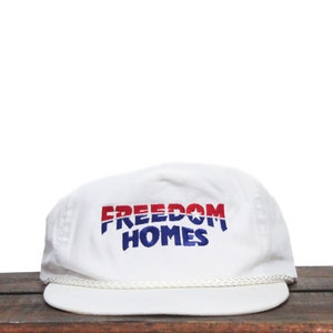 May include: White baseball cap with a red, white and blue embroidered logo that says "FREEDOM HOMES".