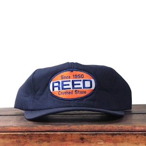 May include: Navy blue baseball cap with a curved brim. The cap features an oval patch with the text "REED Crushed Stone Since 1950" in white and orange. The cap is sitting on a wooden surface.