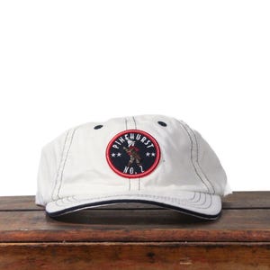 May include: White baseball cap with a black brim and stitching. The cap features a circular patch with the text "PINEHURST NO. 1" and an illustration of a golfer in a red, white, and blue design.