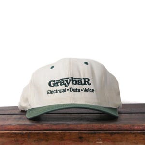 May include: A cream-colored baseball cap with a green brim and accents. The cap features the text "Graybar Electrical Data Voice" in dark green lettering. The hat is sitting on a wooden surface.