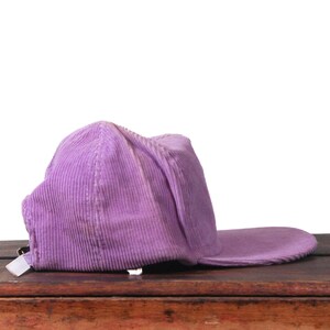 May include: A lavender corduroy baseball cap with a curved brim. The cap is made of a ribbed fabric and has a white tag on the back. The hat is resting on a wooden surface.