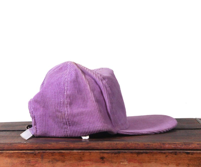 May include: A lavender corduroy baseball cap with a curved brim. The cap is made of a ribbed fabric and has a white tag on the back. The hat is resting on a wooden surface.