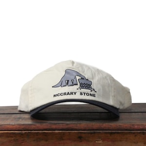 May include: Cream-colored baseball cap with a dark blue brim. The cap features a gray graphic of a stylized animal and the text "MCCRARY STONE" in black. The cap is sitting on a wooden surface.
