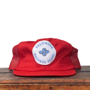 May include: A red trucker hat with a white patch featuring the text "BALTIMORE BUSINESS FORMS" in a circular design. The hat has a mesh back and a curved brim, sitting on a wooden surface.