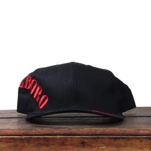 Peut inclure: Casquette de baseball noire avec le mot "BORO" brodé en rouge sur le côté. La casquette a une visière plate et une couronne structurée. La casquette est posée sur une surface en bois.
