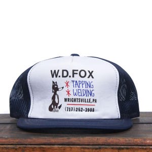 Può includere: Cappellino da camionista bianco e blu con un'illustrazione di volpe in bianco e nero e il testo "W.D. FOX *TAPPING *WELDING WRIGHTSVILLE, PA (717) 252-3908"