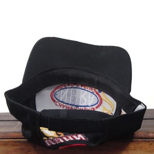 May include: Black baseball cap with a curved brim. The interior features a white patch with blue and red text. The underside of the brim has a colorful design. The cap is resting on a wooden surface.