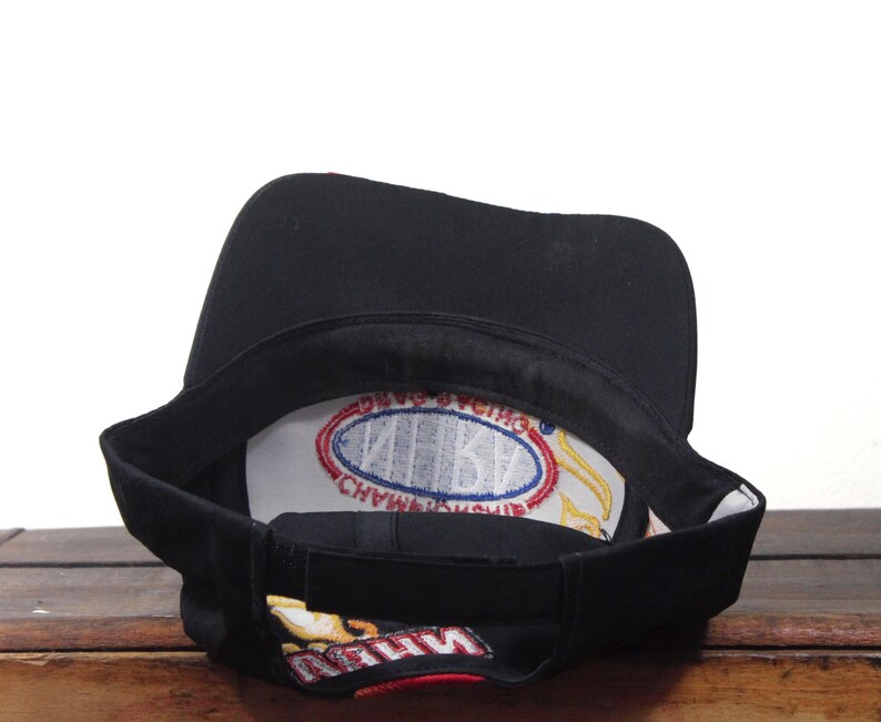 May include: Black baseball cap with a curved brim. The interior features a white patch with blue and red text. The underside of the brim has a colorful design. The cap is resting on a wooden surface.