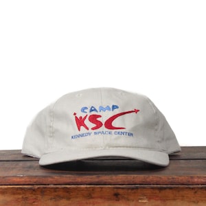 May include: A light gray baseball cap with the text "CAMP KSC KENNEDY SPACE CENTER" embroidered in red and blue. The cap is sitting on a wooden surface.