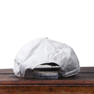 Puede incluir: Gorra de b&eacute;isbol blanca con visera curva, vista desde atr&aacute;s. La gorra est&aacute; hecha de una tela ligeramente texturizada y tiene una correa ajustable. El texto "THERE IS NO FINISH LINE" es visible en la correa.