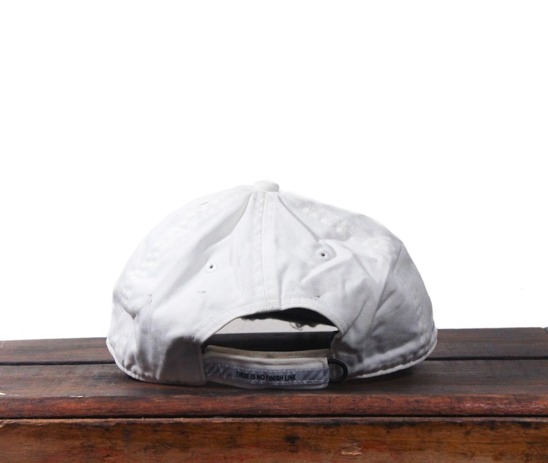 Puede incluir: Gorra de b&eacute;isbol blanca con visera curva, vista desde atr&aacute;s. La gorra est&aacute; hecha de una tela ligeramente texturizada y tiene una correa ajustable. El texto "THERE IS NO FINISH LINE" es visible en la correa.