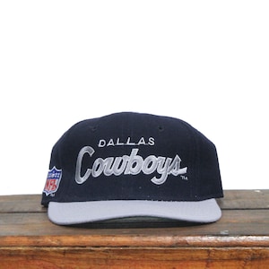 Op de afbeelding: Een marineblauwe en grijze Dallas Cowboys baseballcap. Het woord "DALLAS" is geborduurd boven het woord "Cowboys" in een cursief lettertype. Het NFL-logo staat op de zijkant.