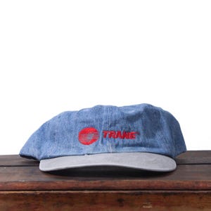 May include: A blue denim baseball cap with a gray brim. The cap features the red word "TRANE" and a red circular logo. The hat is sitting on a wooden surface.