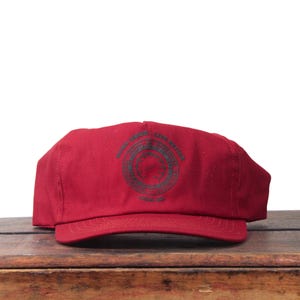 Può includere: Cappellino da baseball rosso con visiera curva. Il cappellino presenta un emblema circolare verde scuro con testo. Il cappellino è appoggiato su una superficie di legno.