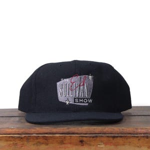 Peut inclure: Casquette de baseball noire avec une visière plate. Le chapeau porte un logo brodé "The Ed Sullivan Show" en argent et rouge. Une étoile est brodée sous le nom de l'émission. Le chapeau est posé sur une surface en bois.
