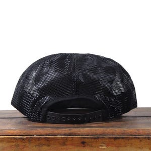 May include: Black trucker hat with a mesh back and a curved brim. The hat features a snapback closure for adjustable sizing. The hat is sitting on a wooden surface.