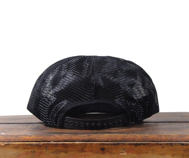 May include: Black trucker hat with a mesh back and a curved brim. The hat features a snapback closure for adjustable sizing. The hat is sitting on a wooden surface.