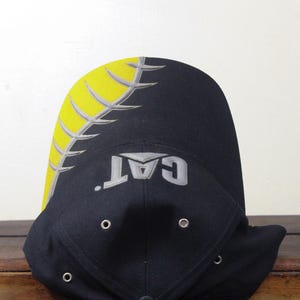 May include: Black baseball cap with a yellow and gray design on the top and the word "CAT" in gray. The cap has a curved brim and eyelets.