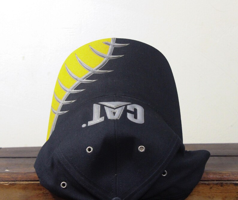 May include: Black baseball cap with a yellow and gray design on the top and the word "CAT" in gray. The cap has a curved brim and eyelets.