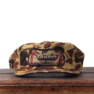 Puede incluir: Gorra de béisbol de camuflaje en tonos marrón y beige. La gorra tiene un parche rectangular con el texto "LEVI GARRETT CHEWING TOBACCO" en marrón y rojo. La gorra está sobre una superficie de madera.