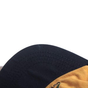 K&ouml;nnte beinhalten: Eine Baseballkappe mit schwarzem Schirm und gelber Krone. Die Unterseite des Schirms ist schwarz. Die Kappe wird von einer Hand gehalten.