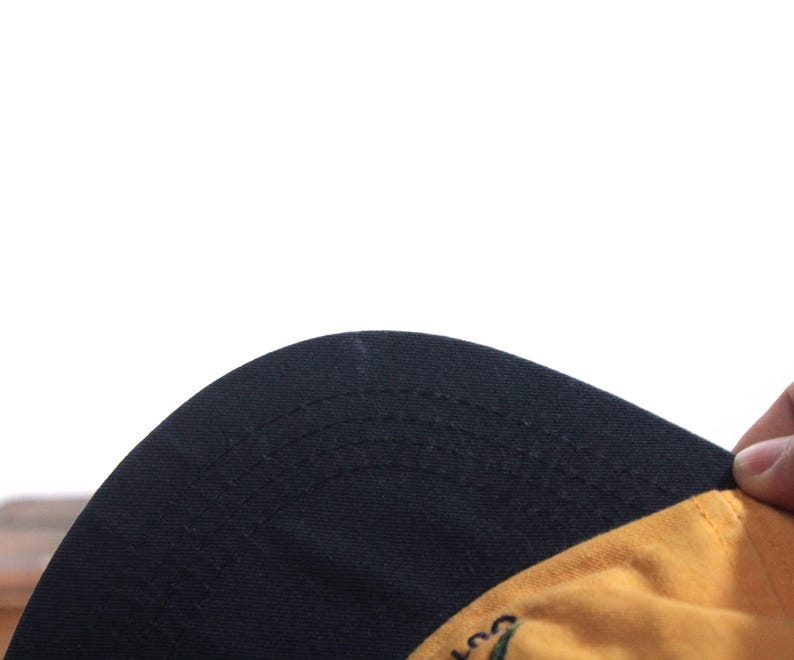 K&ouml;nnte beinhalten: Eine Baseballkappe mit schwarzem Schirm und gelber Krone. Die Unterseite des Schirms ist schwarz. Die Kappe wird von einer Hand gehalten.