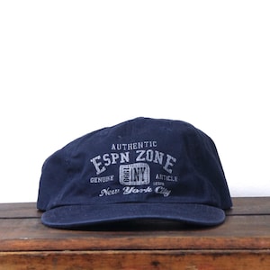 May include: A navy blue baseball cap with the text "AUTHENTIC ESPN ZONE GENUINE ARTICLE 1999 NY New York City" in white. The cap is made of a soft material and has a curved brim.