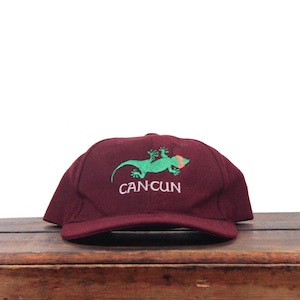 May include: A maroon baseball cap featuring an embroidered green gecko wearing orange sunglasses above the white text CANCUN. The cap has a structured front panel, a curved brim, and a classic six-panel design with a button on the top crown.