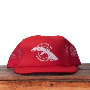 May include: A red baseball cap with a white graphic and text. The cap features a curved brim and mesh side panels. The graphic includes the words "Joint Task Force" and "Guantanamo Bay, Cuba."